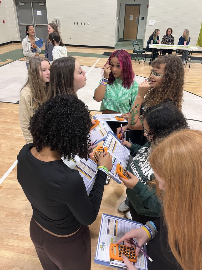 A group of women gathered in a gym, some wearing bracelets, looking at papers and using calculators.