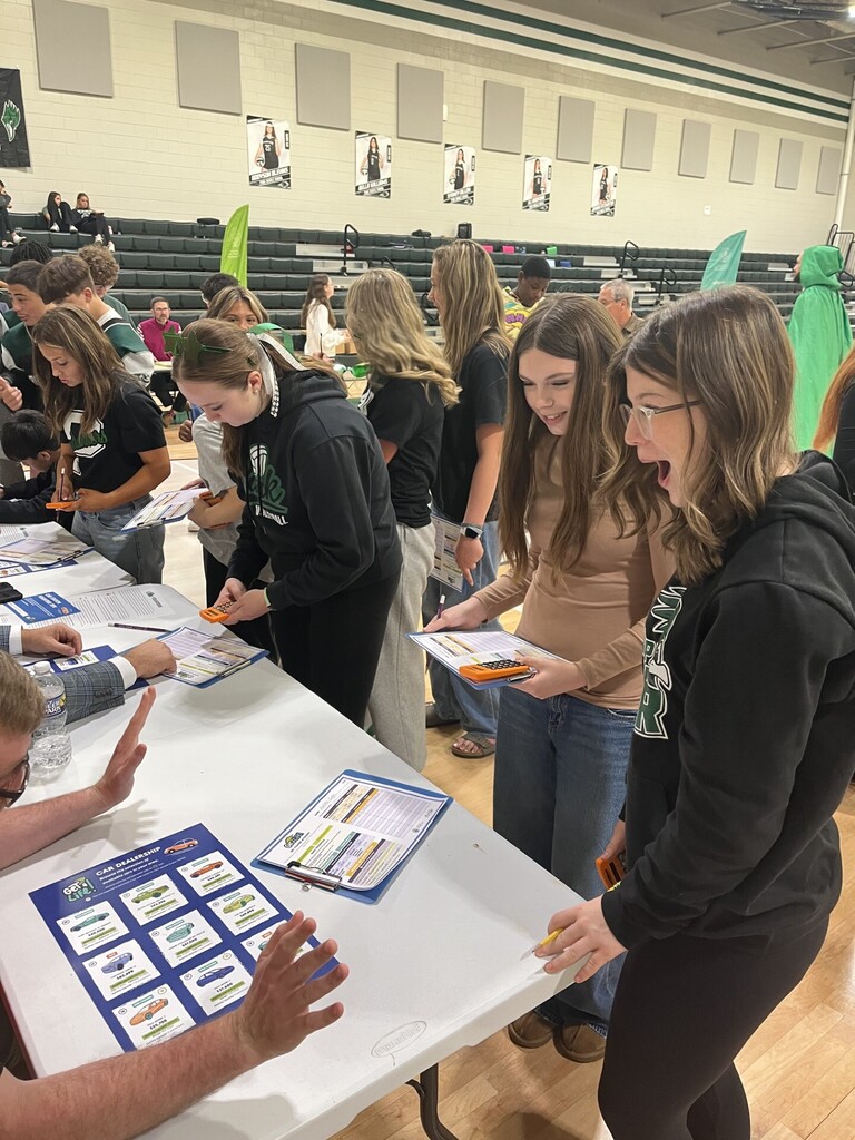 A group of individuals in a school gymnasium, standing around tables with papers and pamphlets.