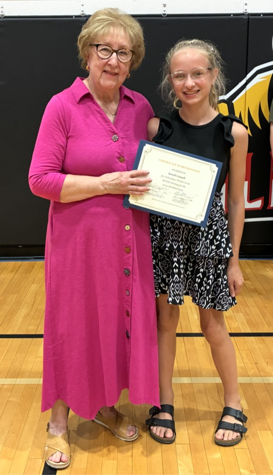 An older woman with glasses and a younger girl with glasses hold a certificate on a basketball court.