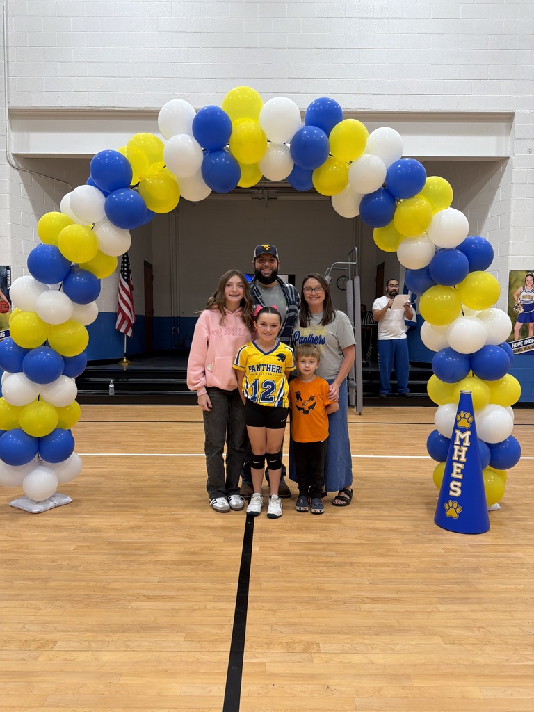 Five people stand in a gym, smiling for a photo. A balloon arch, blue and yellow, stands behind them. A wooden floor and a black line are visible.