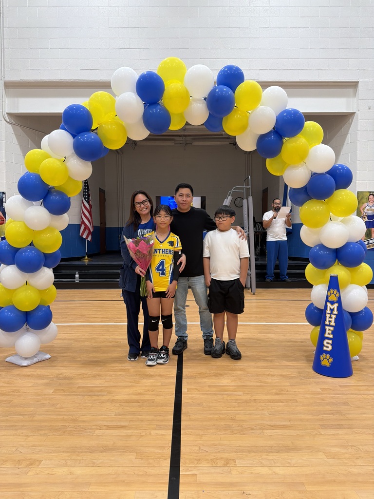 Four people stand under a balloon arch in a gymnasium. A person in the middle wears a sports uniform with a number 4 on it.