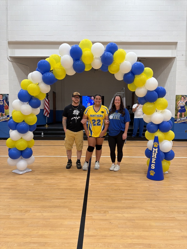 Four individuals stand under a large arch of yellow and white balloons inside a gym. A person wears a jersey with the number 22.