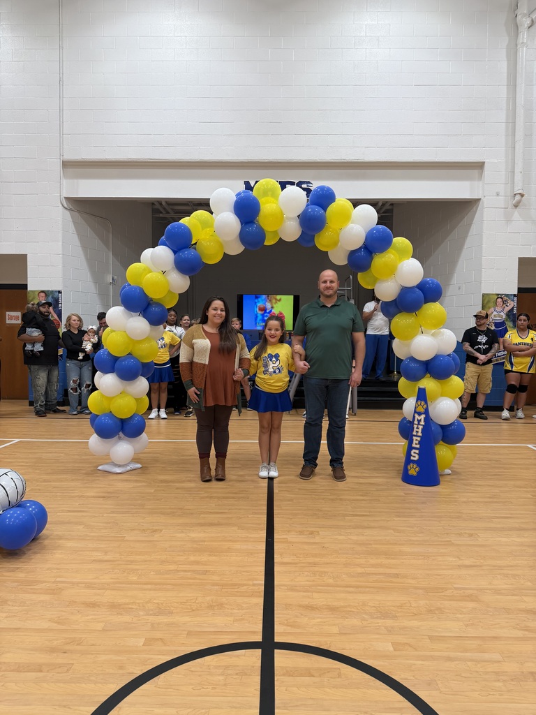 Three people stand under a balloon arch in a gymnasium. A basketball hoop is in the background.