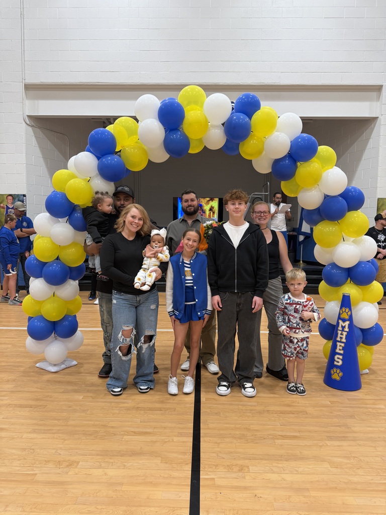 Family and friends pose together behind a balloon arch inside a gym, with blue and yellow balloons.