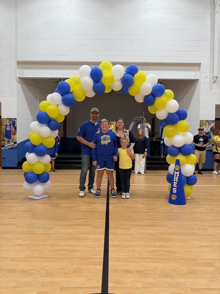 A family of four stands under a balloon arch with blue and white balloons, on a basketball court.