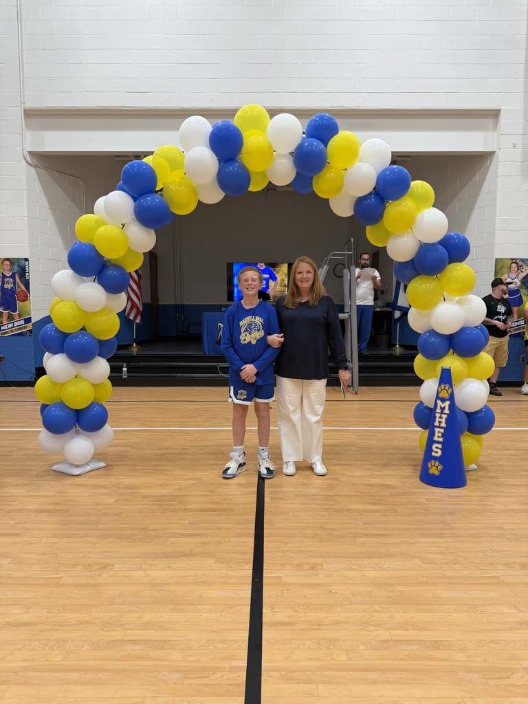Two individuals stand under a colorful balloon arch on a basketball court, with a blue sign in the background.