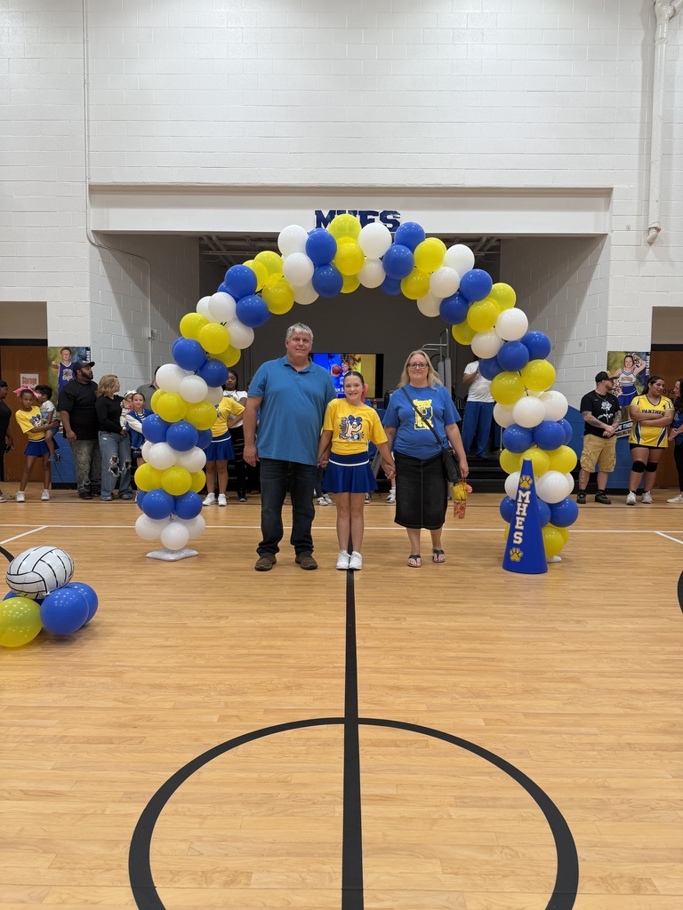 Three people stand beneath a balloon arch on a basketball court. A girl in yellow stands in the middle.