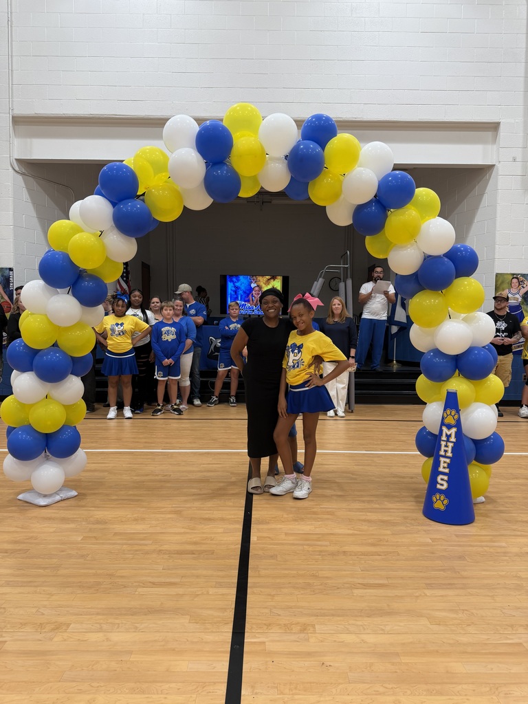 Two individuals stand under a colorful balloon arch on a basketball court, with cheerleaders and onlookers in the background.