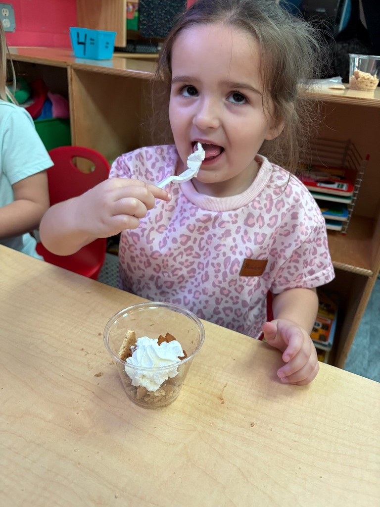 A child eating dessert in a cup at a table. A second child sits behind her.