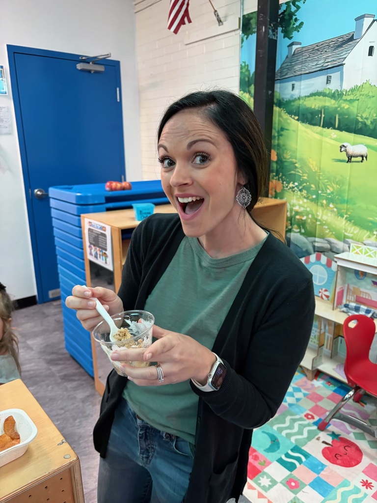 A woman wearing a green shirt and a black cardigan holds a bowl of cereal in a classroom.