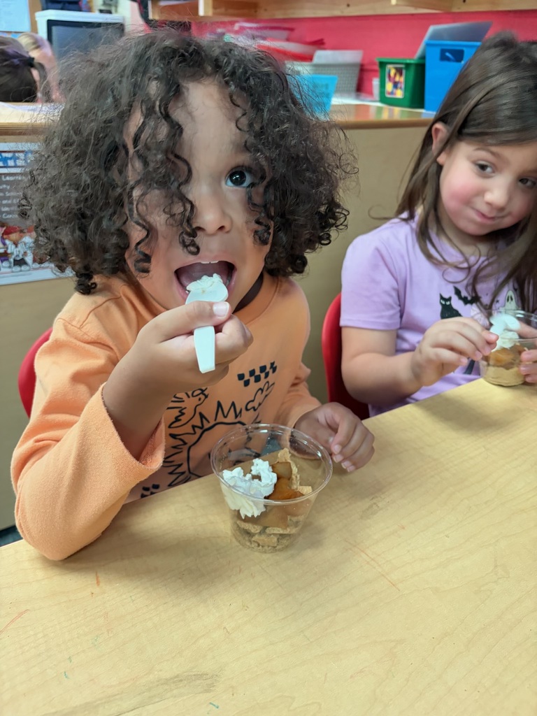 Two children eat yogurt from cups with spoons at a table. One child has curly hair.