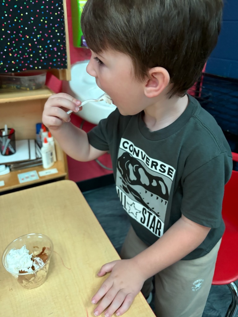 A young boy eats ice cream from a bowl at a table. He wears a black shirt with the word "CONVERSE".