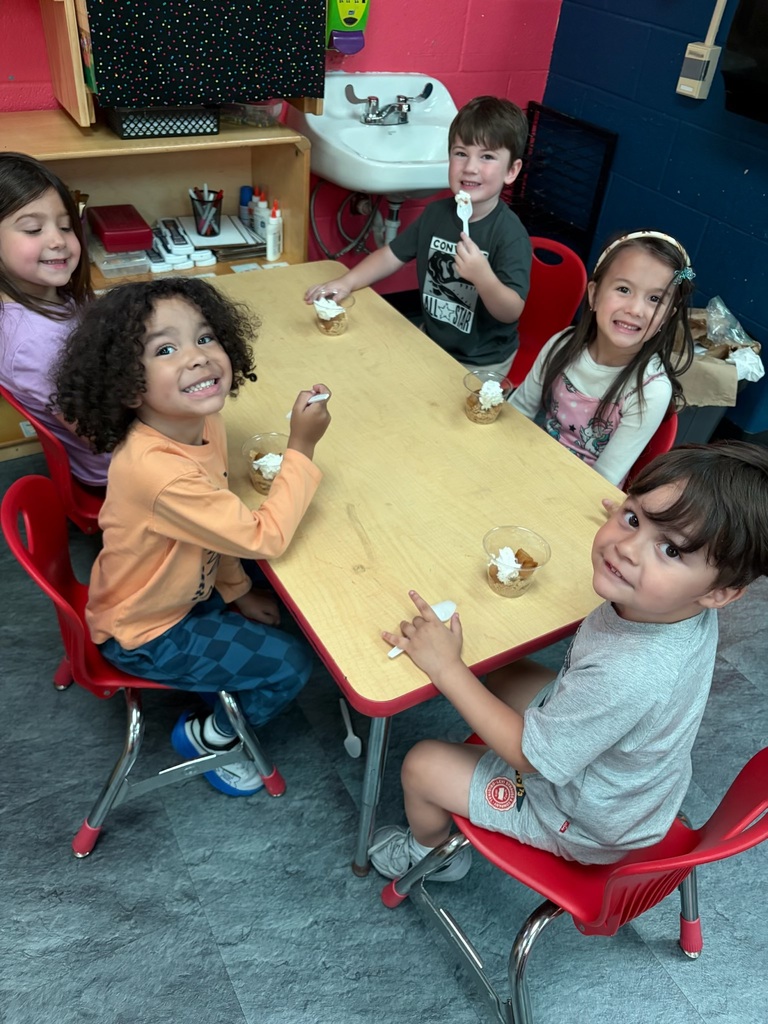 A group of children sit at a table with bowls of food. Behind them is a sink and a wooden shelf.