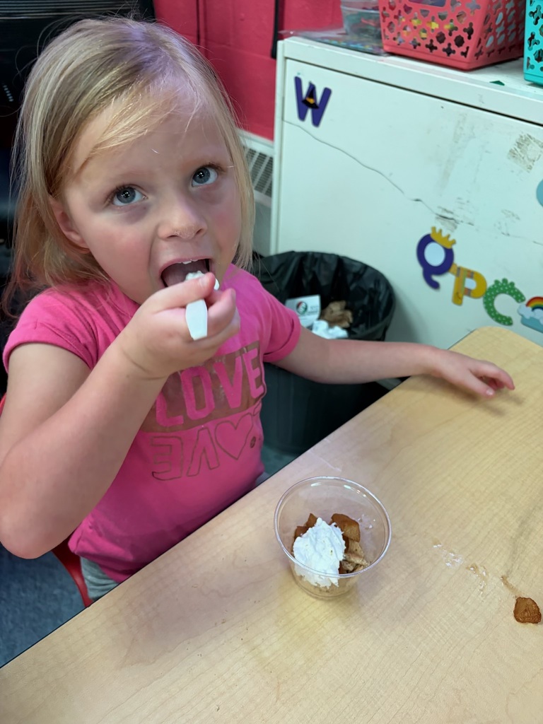 A young girl in a pink shirt eats food from a bowl at a table. A trash can is nearby.