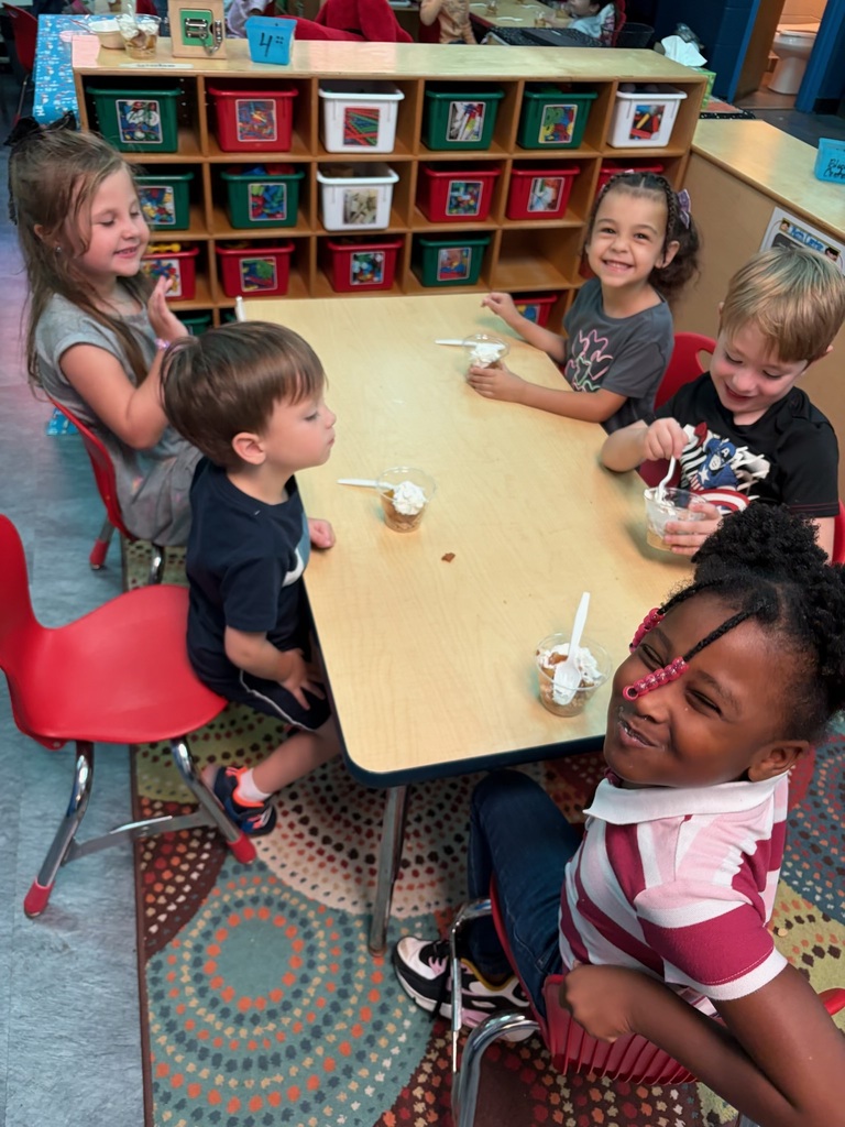 Five children are seated around a table, eating ice cream from bowls. The table is on a rug.