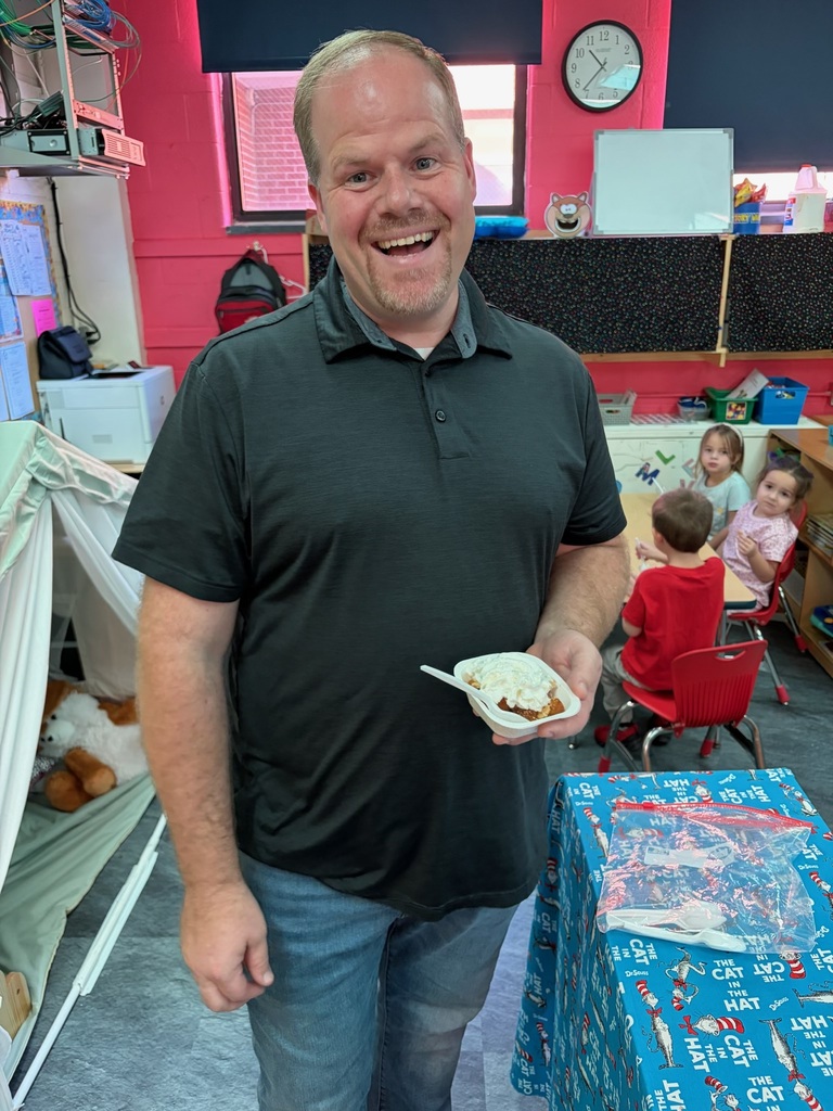 A smiling man in a black shirt holds food in a paper container inside a classroom.