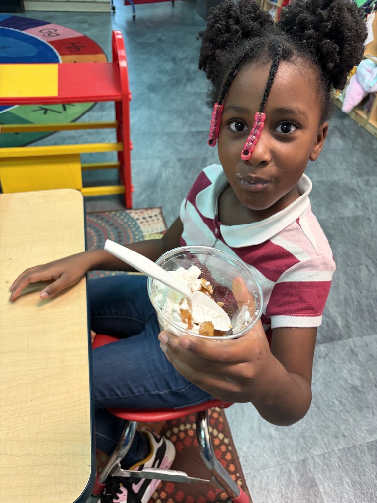 A child eats ice cream from a bowl while seated at a table in a classroom.