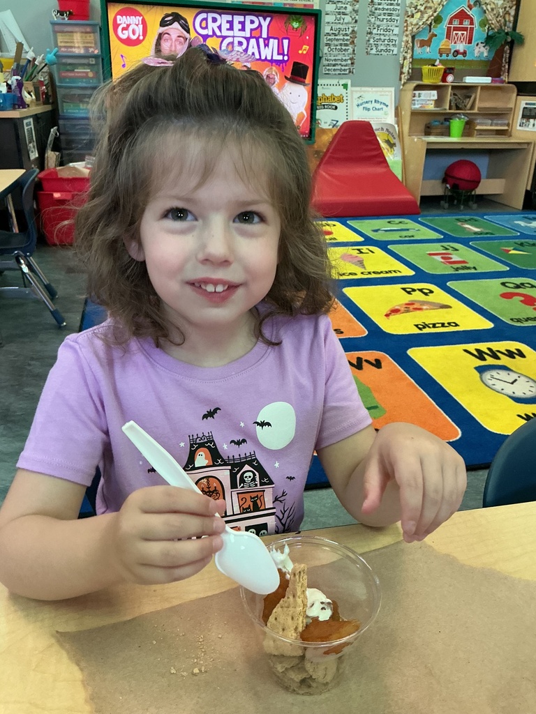A girl sits at a table holding a spoon in a bowl of food. A colorful rug is behind her.