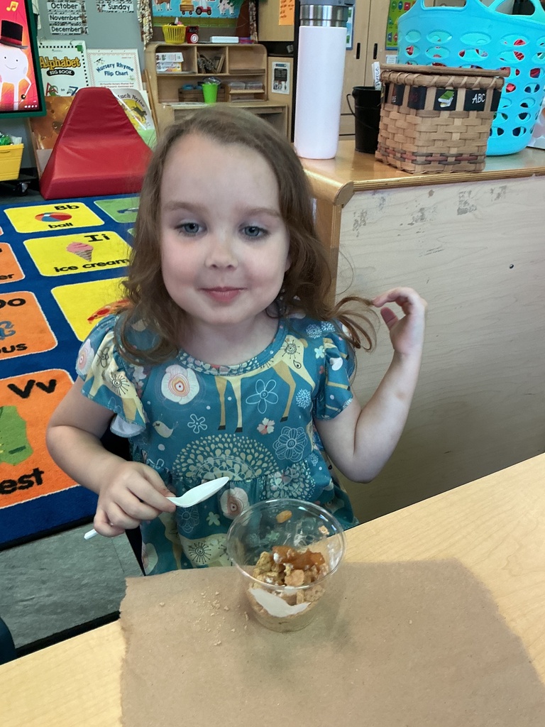A young girl wearing a blue dress sits at a table, holding a spoon in a bowl.