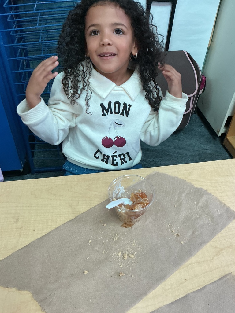 A girl with curly hair sits at a table, smiling. A white shirt says "Mon Cheri." A bowl of food is on the table.