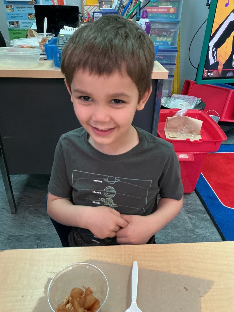 A young boy smiles at the camera, sitting at a table with a bowl of food and a spoon.