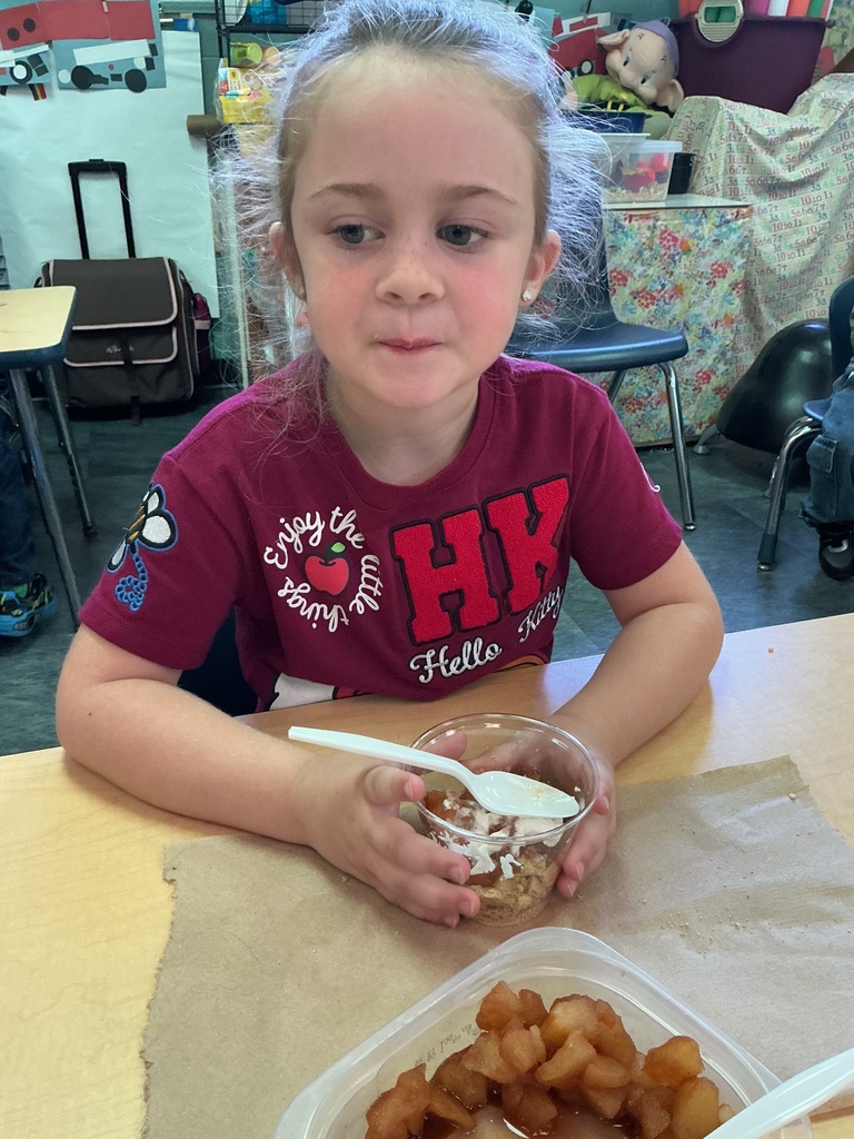 A young girl sits at a table with food in a plastic container and a spoon.