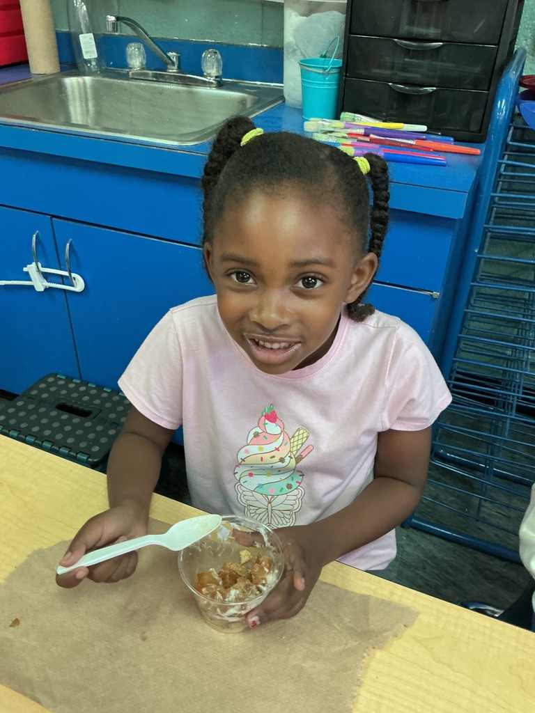 A young girl holds a bowl of food with a spoon. She smiles. A sink, drawers, and a metal rack are behind her.