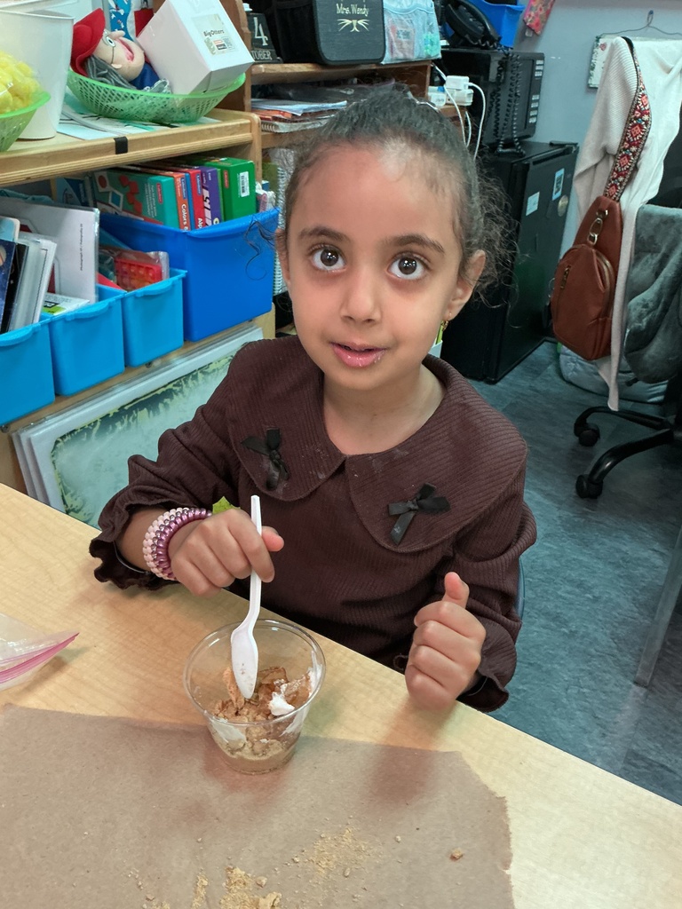 A young girl in a brown shirt, sitting at a table and holding a spoon in a bowl.