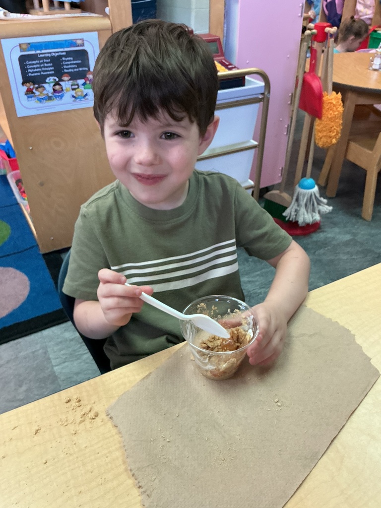 A child in a green shirt sits at a table, holding a spoon in a bowl.