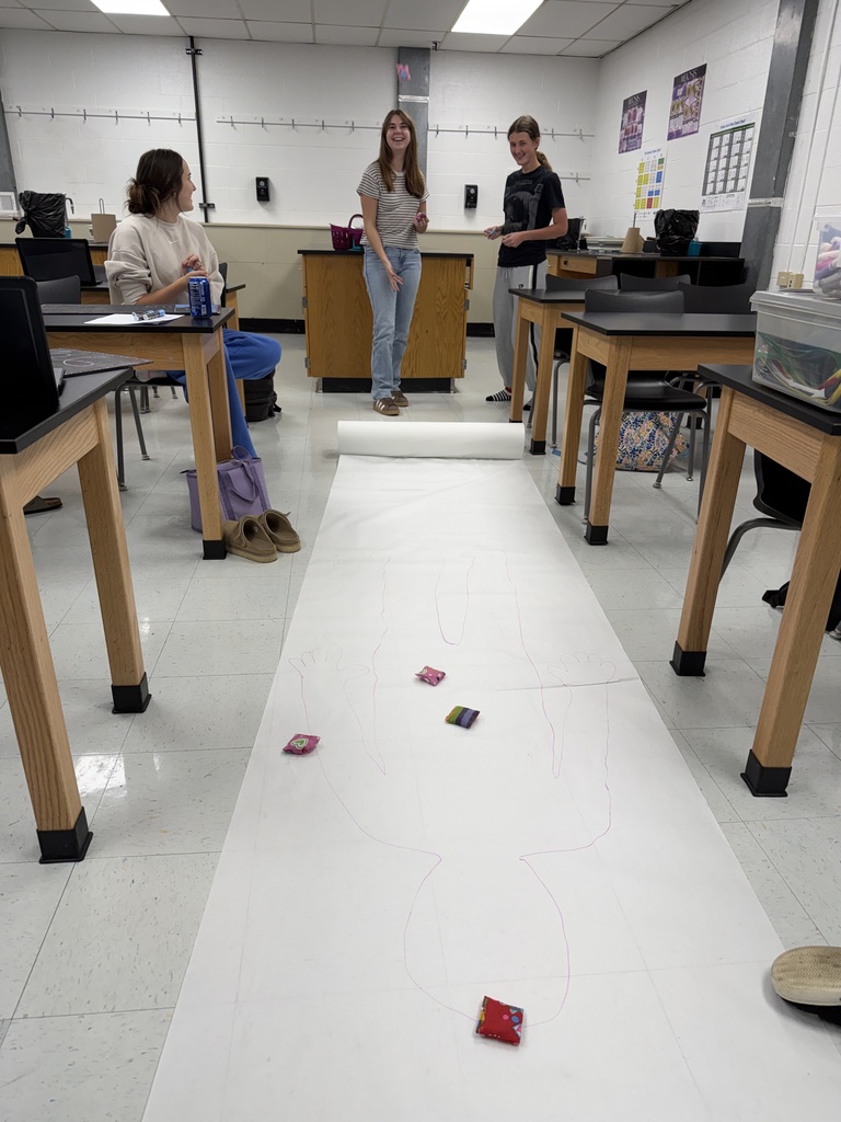 Three students in a classroom, one standing on a white mat, two others standing at a desk.