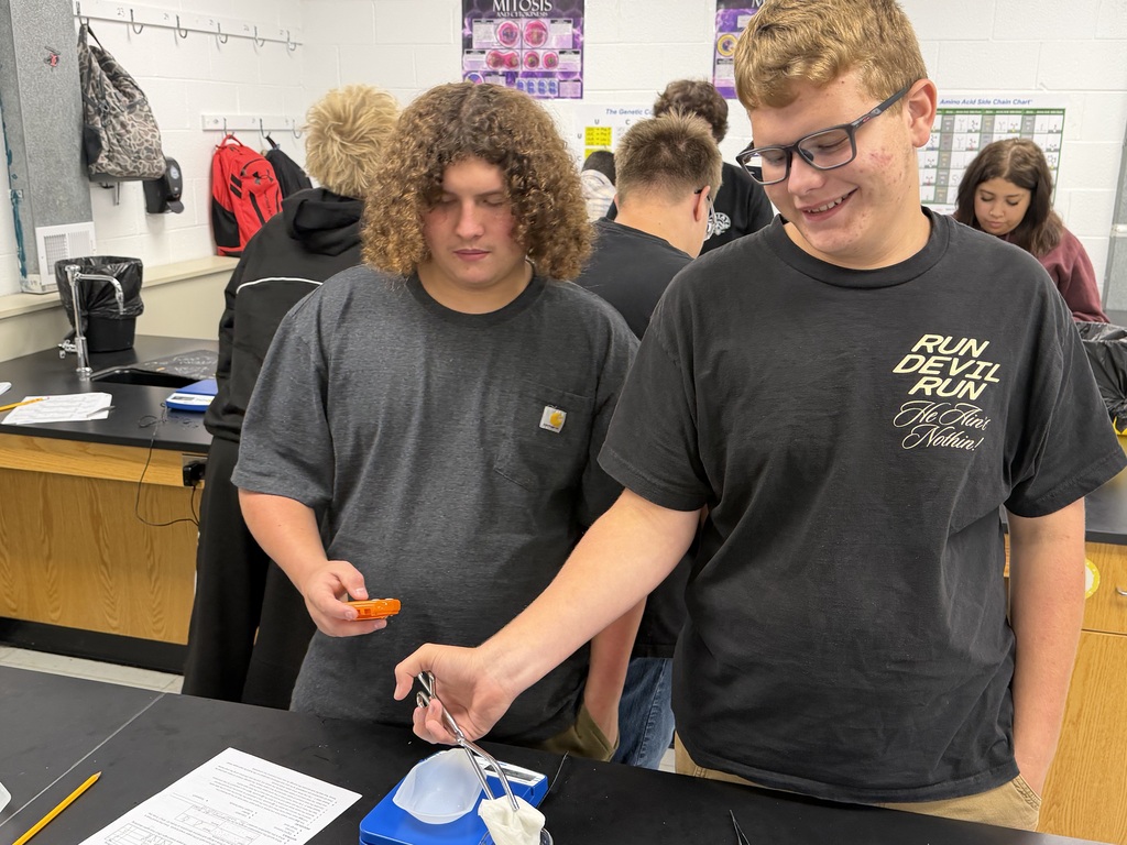 Two students in lab coats in a classroom, one pouring liquid into a cup over a beaker.