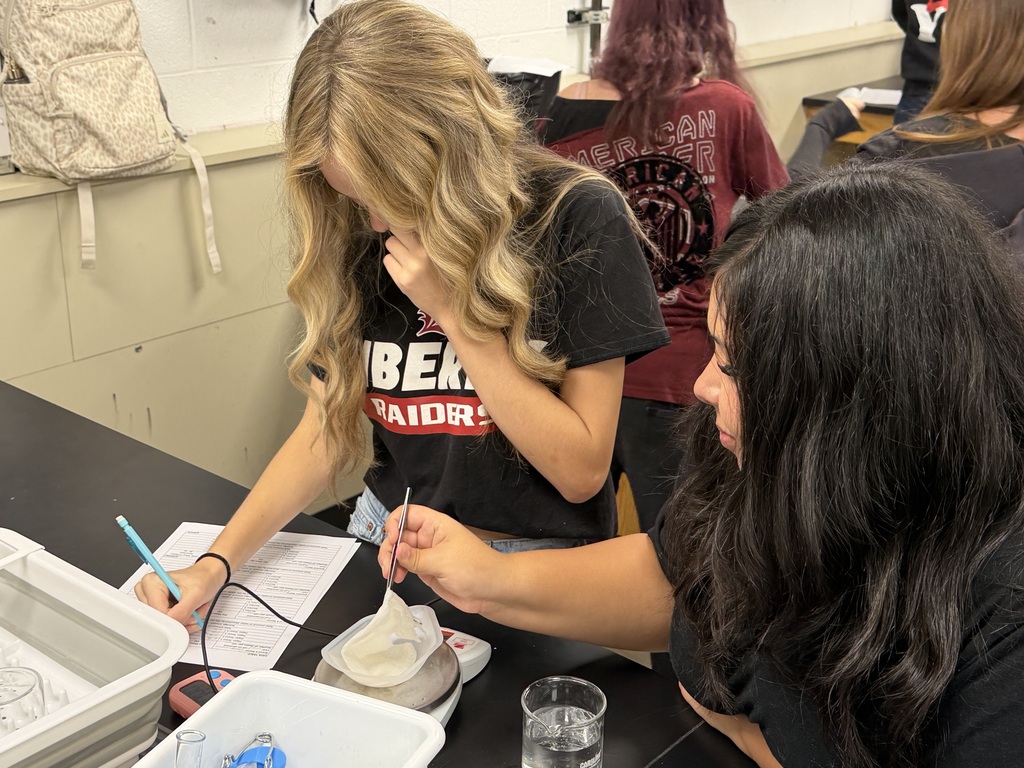 Two students work on a lab project at a table, using a scale, beaker, and spoon.
