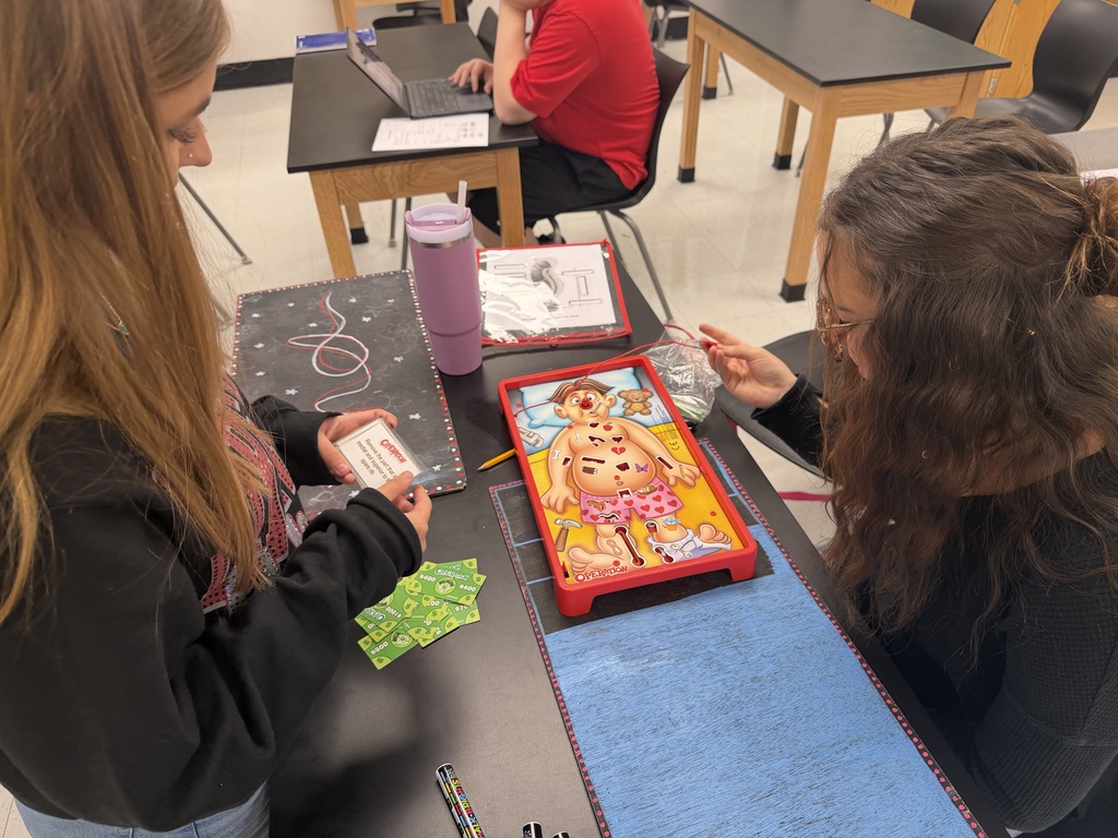 Two girls seated at a table, each holding cards, one with a drawing of a person.