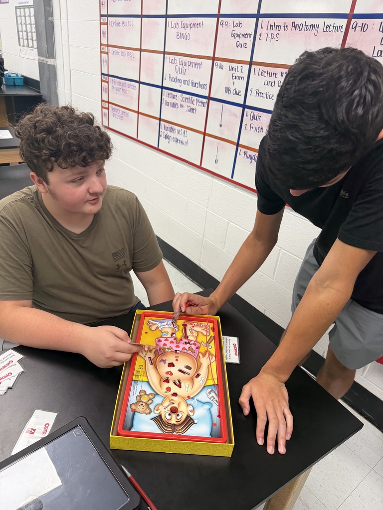 Two students interact with a game on a table, one looking at the game board.