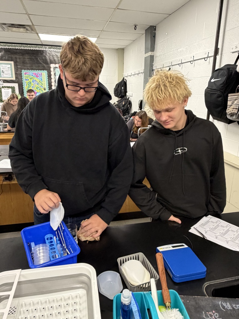 Two students in a classroom look over a lab table with containers, measuring tools, and a white container.