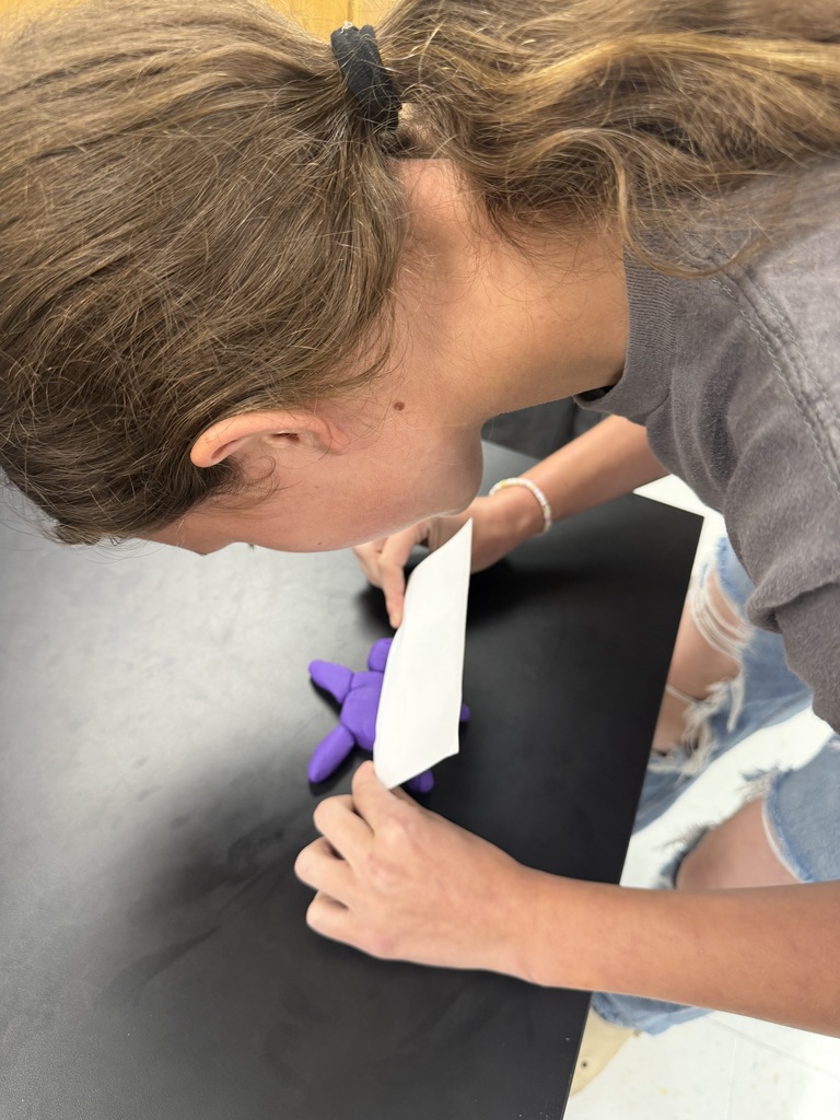 A student with brown hair bent over a black table, holding a white paper and purple object.