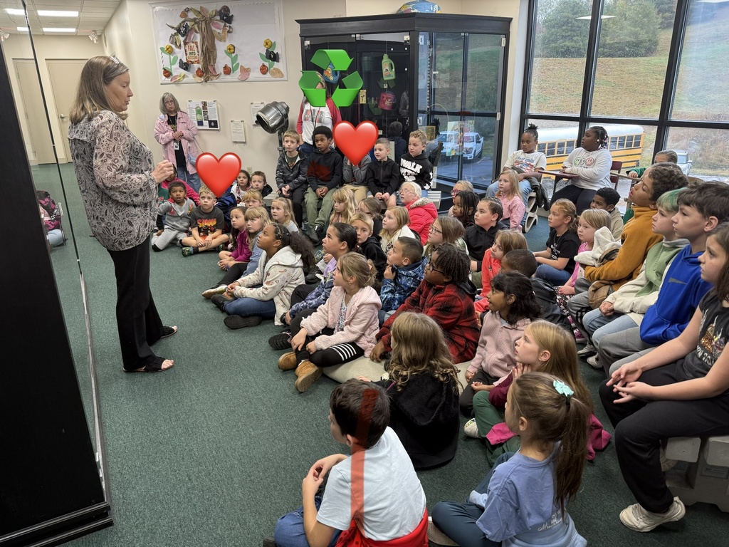 A woman holds red heart balloons in a classroom as students sit on the floor watching.