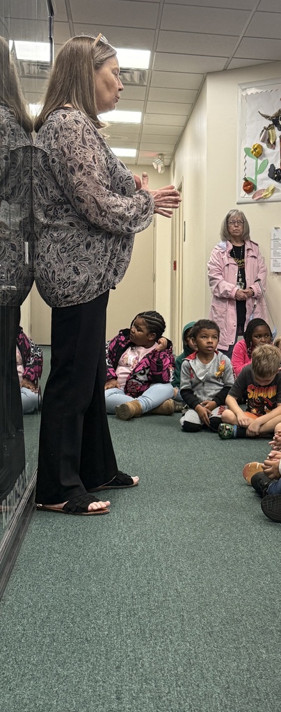 A woman talks to a group of young children sitting on a green floor. A wall displays a paper craft.