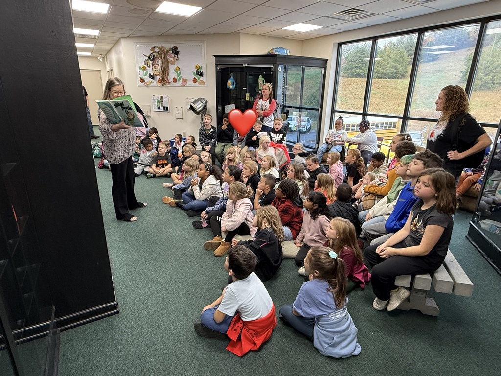 A classroom full of children sitting on the floor listening to a woman read a book.