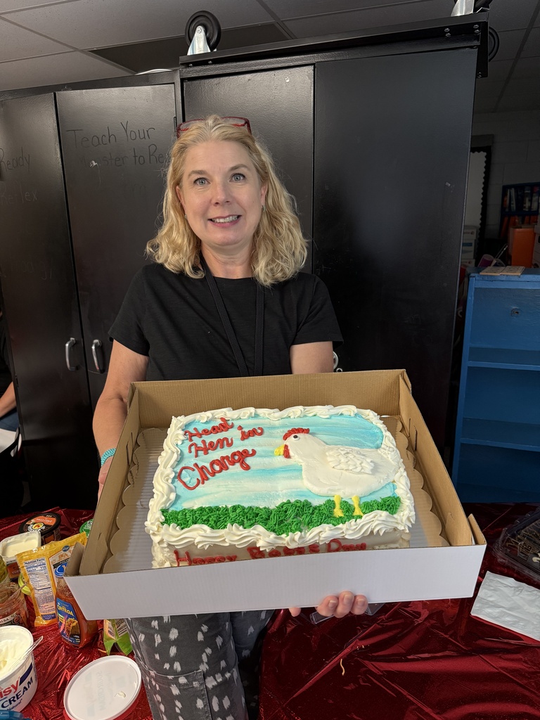 A woman stands holding a box with a cake. The cake has a chicken and words written on it.