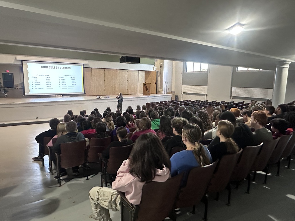 Audience seated in rows, listening to a person standing on a stage with a projection screen