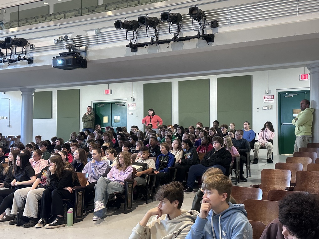 Large classroom with many students seated in rows; two adults at front of room, one standing; overhead lighting.