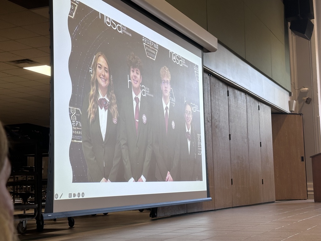 Presentation on projector screen, showing four students in formal attire. Wooden wall panels in background.