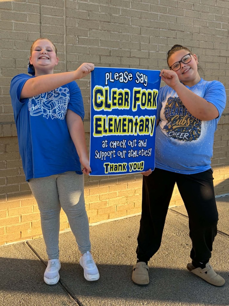 Two students hold up a sign with blue text and a brick wall in the background.