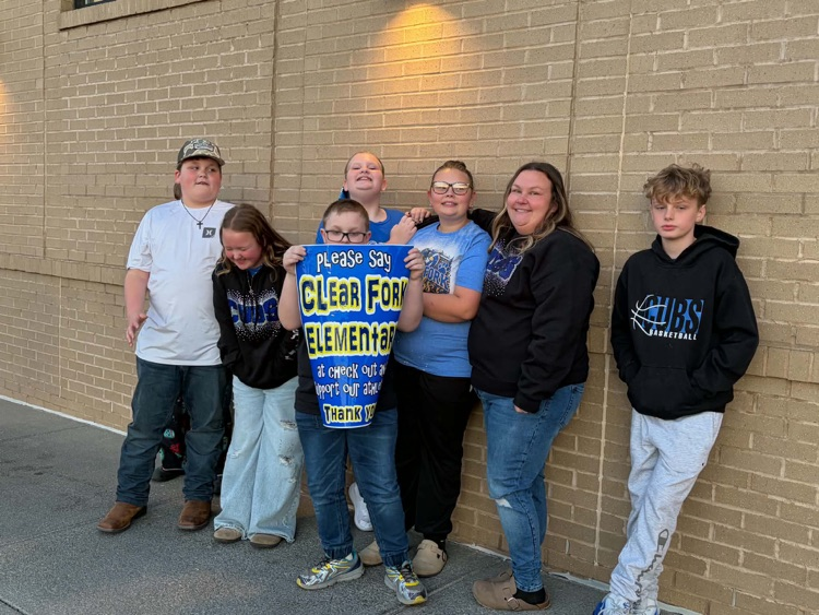 Six students stand against a brick wall, holding a banner with blue writing. They are smiling and posing for a photo.