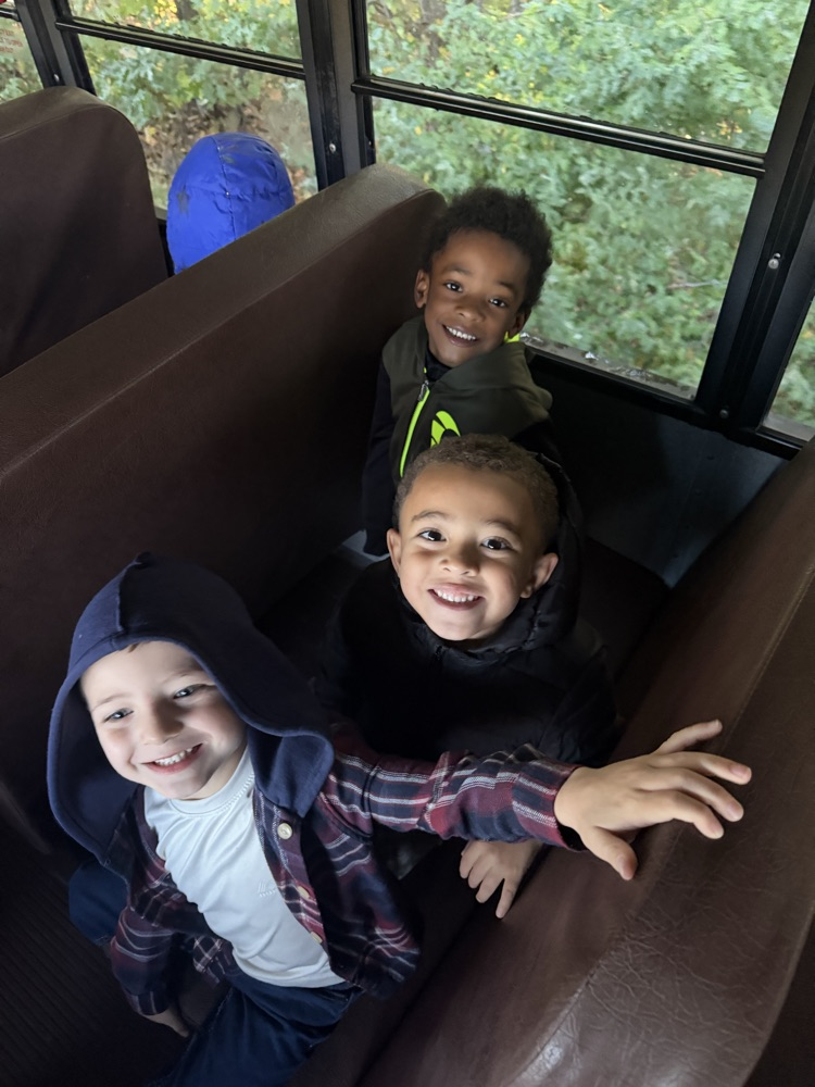 Three children sit together on a brown bus seat. They are smiling. A window behind them shows trees.