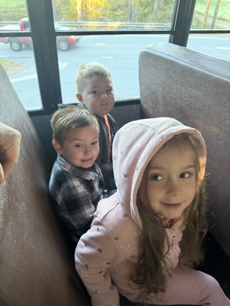 Three children sit together on a bus; one has a hoodie, another has a checkered shirt, and the third wears a black shirt.