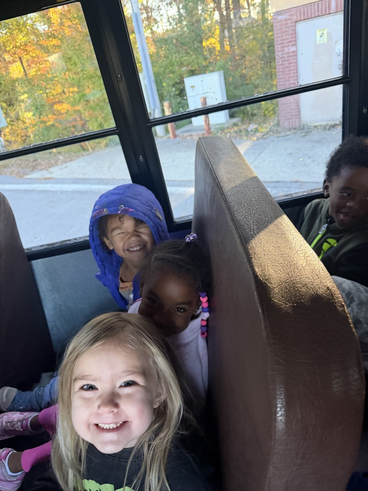 Four children seated on a bus, smiling, with one wearing a blue hoodie, another in a pink shirt, and a brown seat back.
