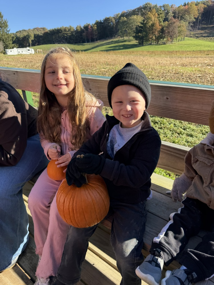 Children in hats holding pumpkins sit on a wooden bench in an outdoor setting.