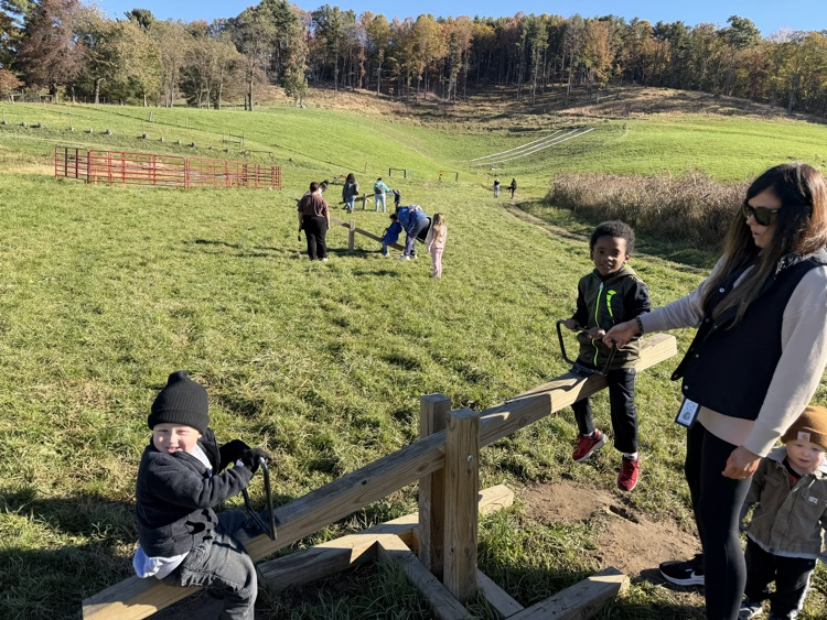 People interact at a playground on a grassy field with trees in the background.